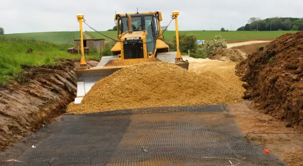 Żółty spychacz Caterpillar przygotowuje teren pod utwardzenie drogi, zasypując wykop piaskiem. Na ziemi rozłożona siatka.