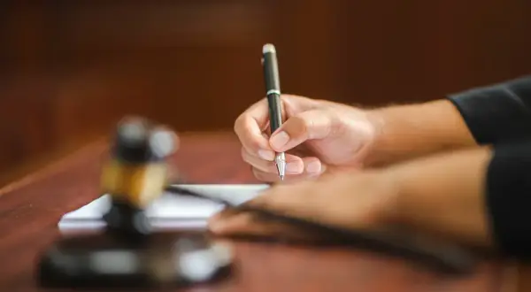 Dłoń referendarza sądowego pisze długopisem na papierze, obok leży młotek sędziowski.