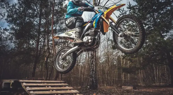 Motocyklista w kasku i goglach skacze na motocyklu enduro nad drewnianą rampą w lesie.