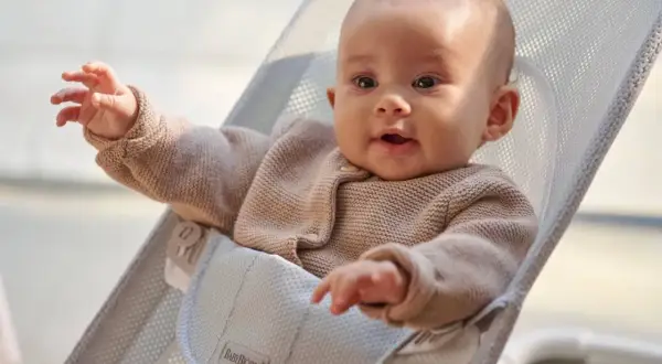 Uśmiechnięte niemowlę w beżowym sweterku siedzi w leżaczku BabyBjörn. Ten leżaczek jest idealny od pierwszych miesięcy życia dziecka.