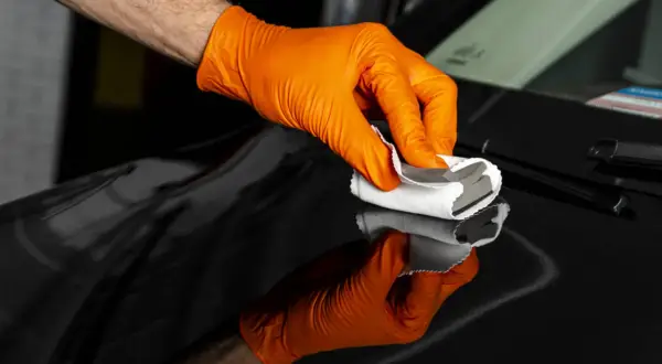 Ręka w pomarańczowej rękawiczce aplikuje na czarną karoserię samochodu produkt do nakładania powłoki ceramicznej.