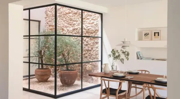 Un comedor moderno con una mesa de madera y sillas de diseño, junto a un patio de luces con olivos en macetas y paredes de piedra.