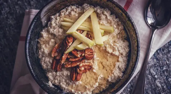 Porridge zubereiten: Cremiger Haferbrei mit Apfelspalten, Pekannüssen und Honig in einer Schüssel, daneben ein Löffel.