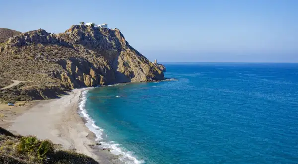 Costa rocosa con una playa de arena dorada y aguas turquesas. En lo alto del acantilado, casas blancas y un faro vigilan la costa. Un lugar idílico para disfrutar del sopalmo playa.