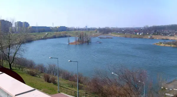 Spokojne jezioro Zatorze otoczone zielenią i zabudowaniami miejskimi. Na wodzie mała wyspa z drzewami.