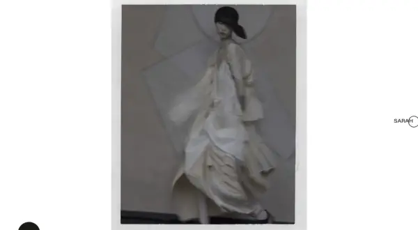 Modelka w zwiewnej, kremowej sukni i czarnym kapeluszu, w stylu Sarah Moon, porusza się z gracją.