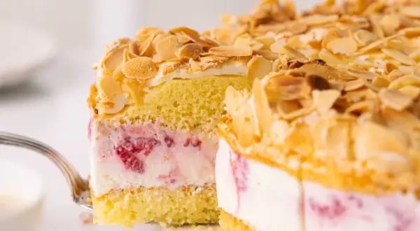 Ein Stück himmelstorte mit himbeeren wird mit einer Gabel herausgehoben. Die Torte ist mit Mandelsplittern bestreut.