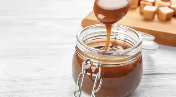 Une cuillère verse un coulis de caramel onctueux dans un bocal en verre. Des bonbons au caramel sont disposés sur une planche en bois en arrière-plan.