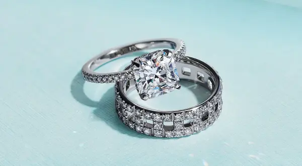Un anillo de compromiso con un gran diamante cuadrado reposa sobre un anillo de matrimonio con incrustaciones de diamantes, ambos de plata.
