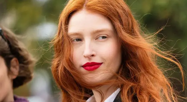 Eine Frau mit leuchtend roten Haaren, die im Wind wehen. Ihr Blick ist nach rechts gerichtet, ihre Lippen sind rot geschminkt. Natur und rote Haare vereinen sich in diesem Porträt.