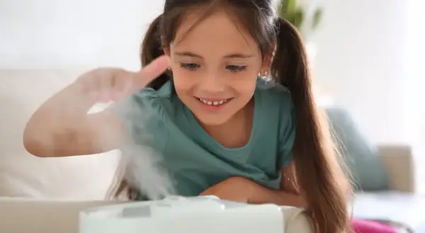 Uśmiechnięta dziewczynka bawi się mgiełką z nawilżacza powietrza dla niemowląt, tworząc magiczną atmosferę w pokoju.