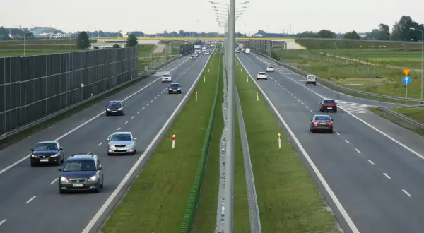 Samochody poruszają się po wielopasmowej trasie szybkiego ruchu, otoczonej zielenią i ekranami akustycznymi.
