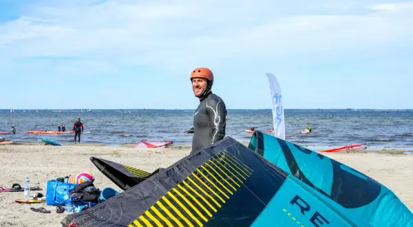 Mężczyzna w kasku i piance na plaży, leżące latawce do kitesurfingu, morze i inni sportowcy.