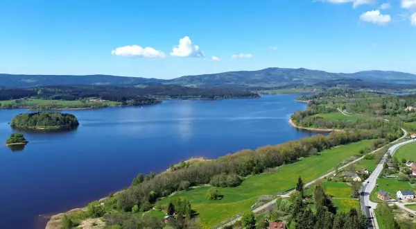 Weite Sicht auf den Lipno Stausee mit bewaldeten Hügeln, grünen Wiesen und kleinen Häusern am Ufer.