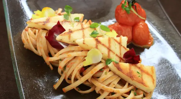 Makaron z halloumi, grillowane plastry sera, pomidorki koktajlowe i listki ziół na szklanym talerzu.