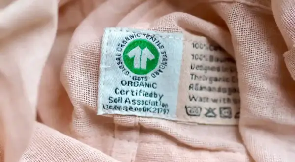 Metka z symbolem GOTS Organic na różowej tkaninie. Informuje o ekologicznym pochodzeniu materiału i zaleca delikatne pranie, bez suszenia w suszarce bębnowej.