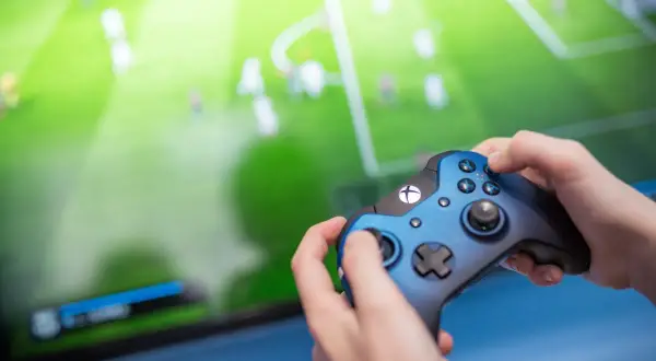 Ręce trzymające niebieski kontroler Xbox, grające w grę piłkarską. Ekran telewizora pokazuje zielone boisko z zawodnikami, tworząc efektowną teselację.