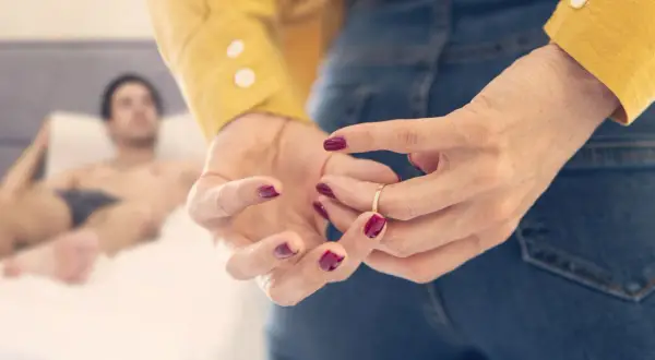 Vrouw met rode nagellak speelt met haar trouwring, terwijl een man op de achtergrond ligt. De vraag "hoe vaak komt een affaire uit" hangt in de lucht.