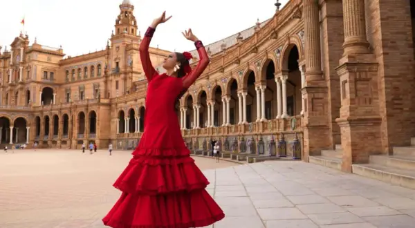Bailarina con vestido rojo voluminoso, brazos en alto, ejecutando sevillanas lentas frente a un edificio histórico con arcos.