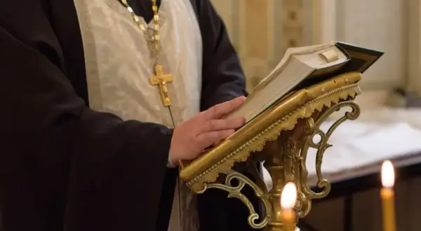 Duchowny w szatach, z krzyżem na szyi, czyta księgę podczas nabożeństwa prawosławnego.