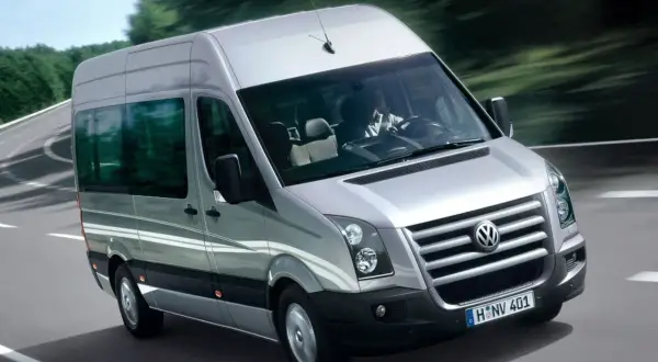 Srebrny Volkswagen Crafter jedzie po krętej drodze wśród drzew. Kierowca w białej koszuli prowadzi auto.