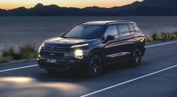 Ciemny Mitsubishi Outlander PHEV jedzie drogą o zachodzie słońca, z górami i jeziorem w tle.