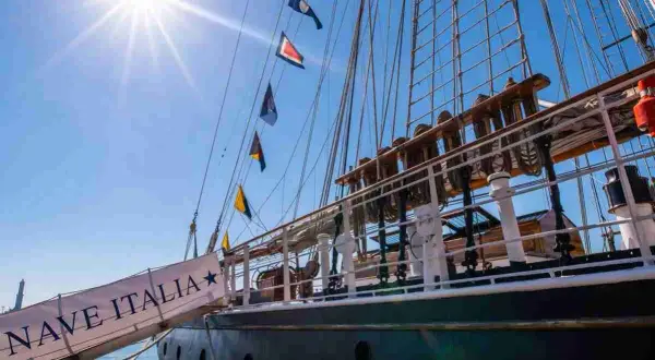 La nave Italia, con le sue vele spiegate e le bandiere colorate, si staglia contro il cielo azzurro.