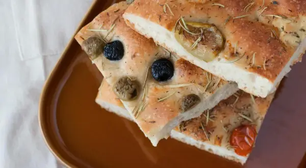 Focaccia Pomodoro mit Oliven und Rosmarin auf brauner Platte.