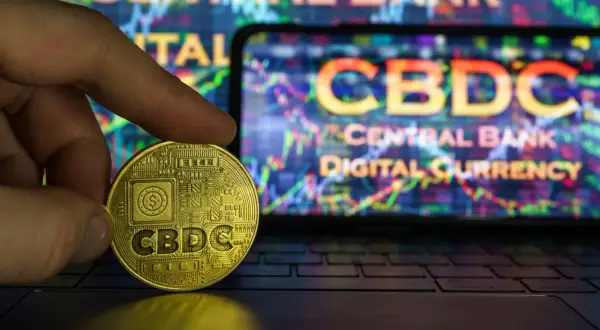 Złota moneta z napisem CBDC w dłoni na tle ekranu z wykresem i napisem "Central Bank Digital Currency". To przyszłość finansów, ale cbdc co to?