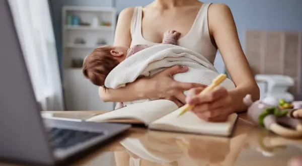 Kobieta z dzieckiem na rękach notuje, zastanawiając się, czy macierzyński wlicza się do emerytury.