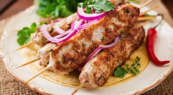 Aksamitny kebab z indyka na talerzu, ozdobiony krążkami czerwonej cebuli i świeżą natką pietruszki. Obok leży pikantna papryczka chili.