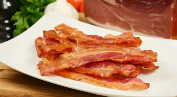 Knusprig gebratener Bacon im Ofen, auf einem weißen Teller gestapelt. Im Hintergrund ein Stück rohes Fleisch, Knoblauch und Petersilie.
