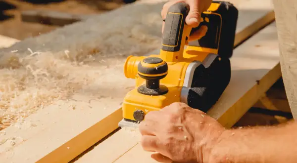 Un artisan utilise une raboteuse électrique pour raboter une porte en bois, produisant des copeaux fins.
