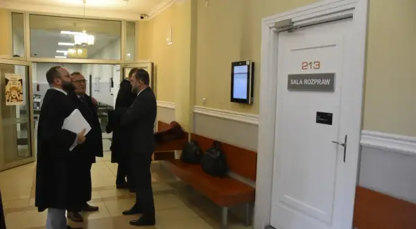 Trzech mężczyzn w togach prawniczych rozmawia przed drzwiami sali rozpraw nr 213.