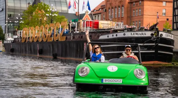 Para wodna w Bydgoszczy: młoda para w zielonym samochodziku-łódce, na wodzie, z napisem "BYDGOSZCZ" na barce w tle. To świetne bydgoszcz atrakcje!