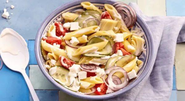 Schüssel mit griechischem Nudelsalat: Penne, Gurken, Tomaten, rote Zwiebeln und Feta in cremiger Soße.