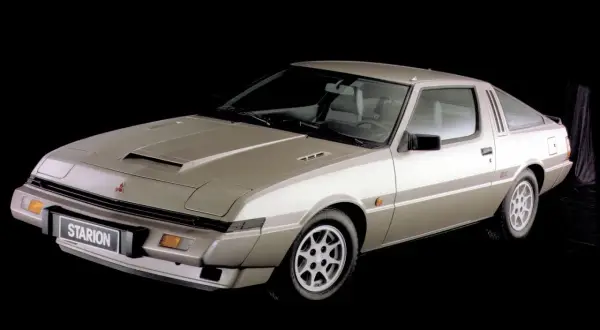 Klasyczny Mitsubishi Starion w kolorze szampańskim, z charakterystycznym wlotem powietrza na masce, prezentuje się na czarnym tle.