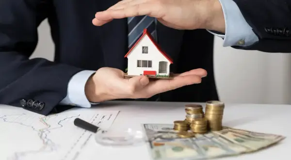 Mani che proteggono una casa modello, con grafici, soldi e monete. L'idea di comprare casa per affittarla è ben rappresentata.