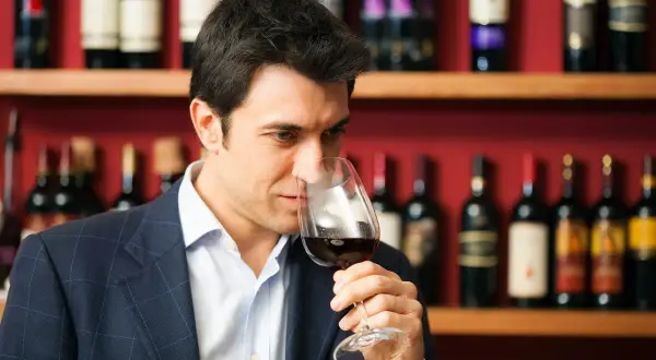 Mężczyzna w garniturze wącha czerwone wino w kieliszku. To degustator wina, który ocenia jego aromat.
