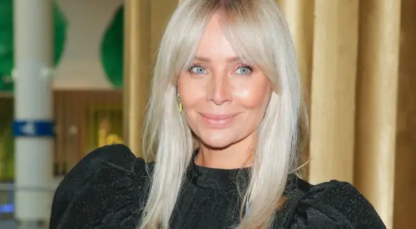 Kobieta z jasnymi włosami i modną fryzurą curtain bangs uśmiecha się do obiektywu.