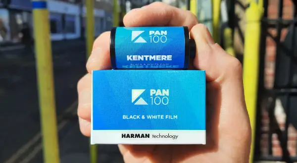 A hand holds a roll and box of Kentmere Pan 100 black and white film.