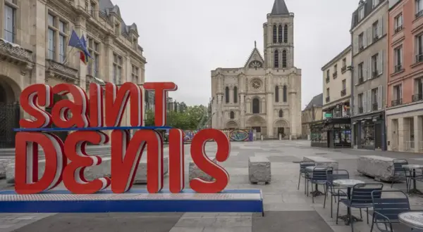 Duże czerwone litery "SAINT DENIS" na tle bazyliki. Choć nazwa sugeruje niebezpieczne miejsca, plac wydaje się spokojny.