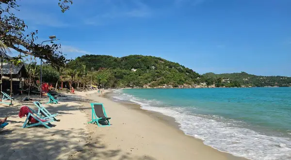 Solig strand på Koh Phangan, Thailand. Turkost vatten, gröna kullar och tomma solstolar väntar på besökare.
