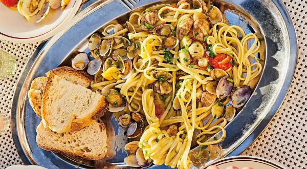 En stor silverbricka med pasta vongole, musslor, körsbärstomater och vitlöksbröd. En hand tar en gaffel med pasta.