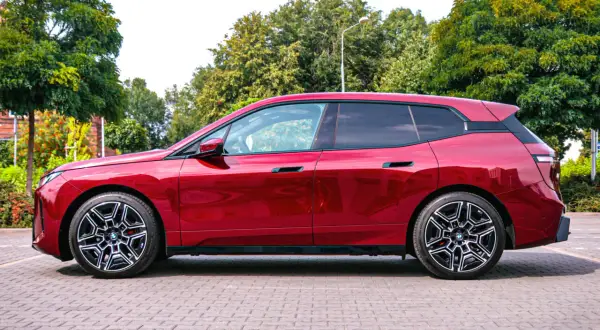 Błyszczący, czerwony BMW iX stoi na brukowanym placu, otoczony zielenią.