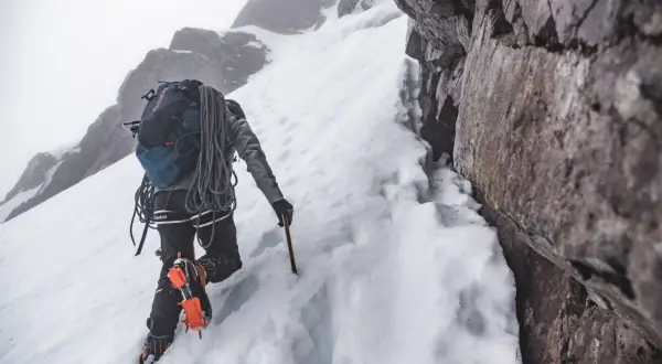 Grimpeur en crampons et piolet, progressant sur une pente enneigée. Un entraînement en altitude exigeant, au cœur d'un paysage rocheux et brumeux.