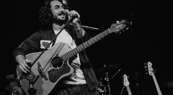 Un postino cantante con chitarra e microfono sul palco, circondato da strumenti musicali.