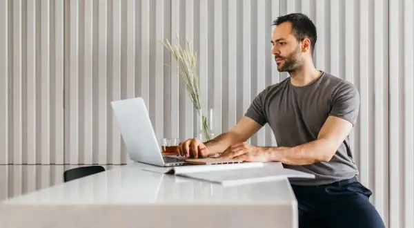 Mężczyzna w szarym t-shircie pracuje na laptopie przy białym stole.