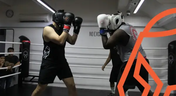 Two boxers spar in a ring, practicing their boxing defense. One boxer wears a black tank top and headgear, the other a grey tank top and white headgear.
