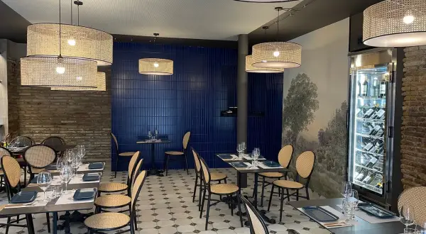 Interior de la brasserie 1885, con mesas puestas, sillas de mimbre y lámparas de ratán. Pared azul y mural de paisaje.